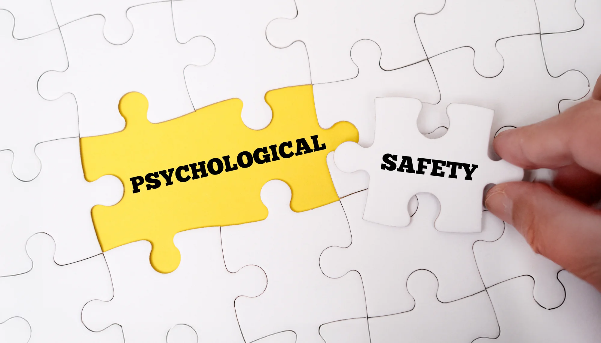 Weiß-gelbes Puzzle mit Schriftzug Psychological Safety, eine Hand hält einen Puzzlestein